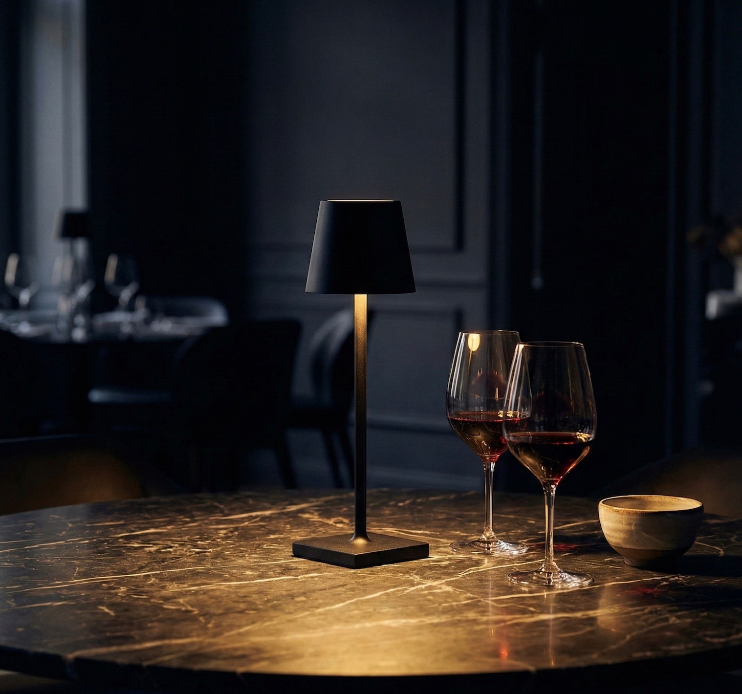 Black table lamp on a marble surface with two wine glasses and a bowl in a dimly lit room.